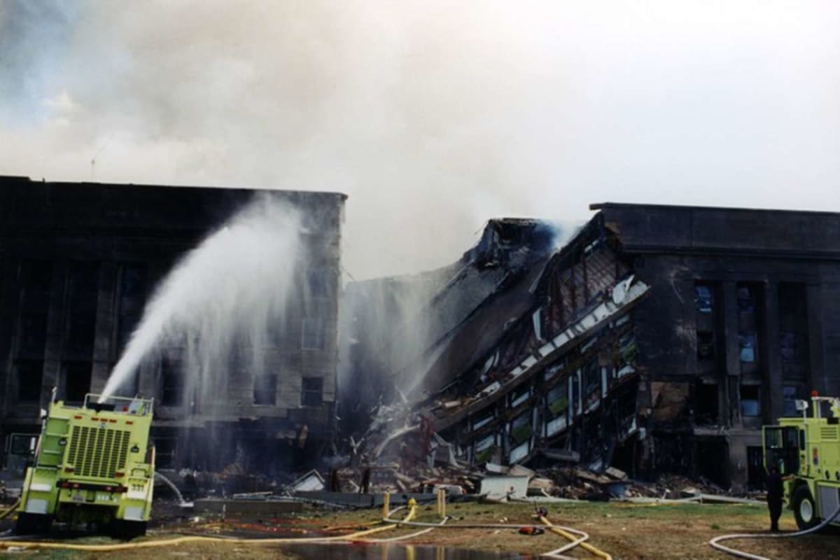 Firefighters spray water on a partially collapsed building with a tilted roof and debris on the ground, two yellow fire engines nearby.