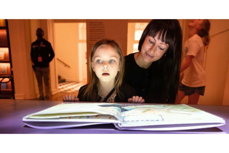 A young girl and a woman lean over an open, oversized book glowing on a purple table in a museum setting.