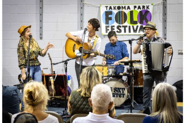 Band performs at Takoma Park Folk Festival; guitarist, singer, drummer, and accordion player on a small stage in front of a banner.