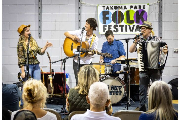 Band performs at Takoma Park Folk Festival; guitarist, singer, drummer, and accordion player on a small stage in front of a banner.