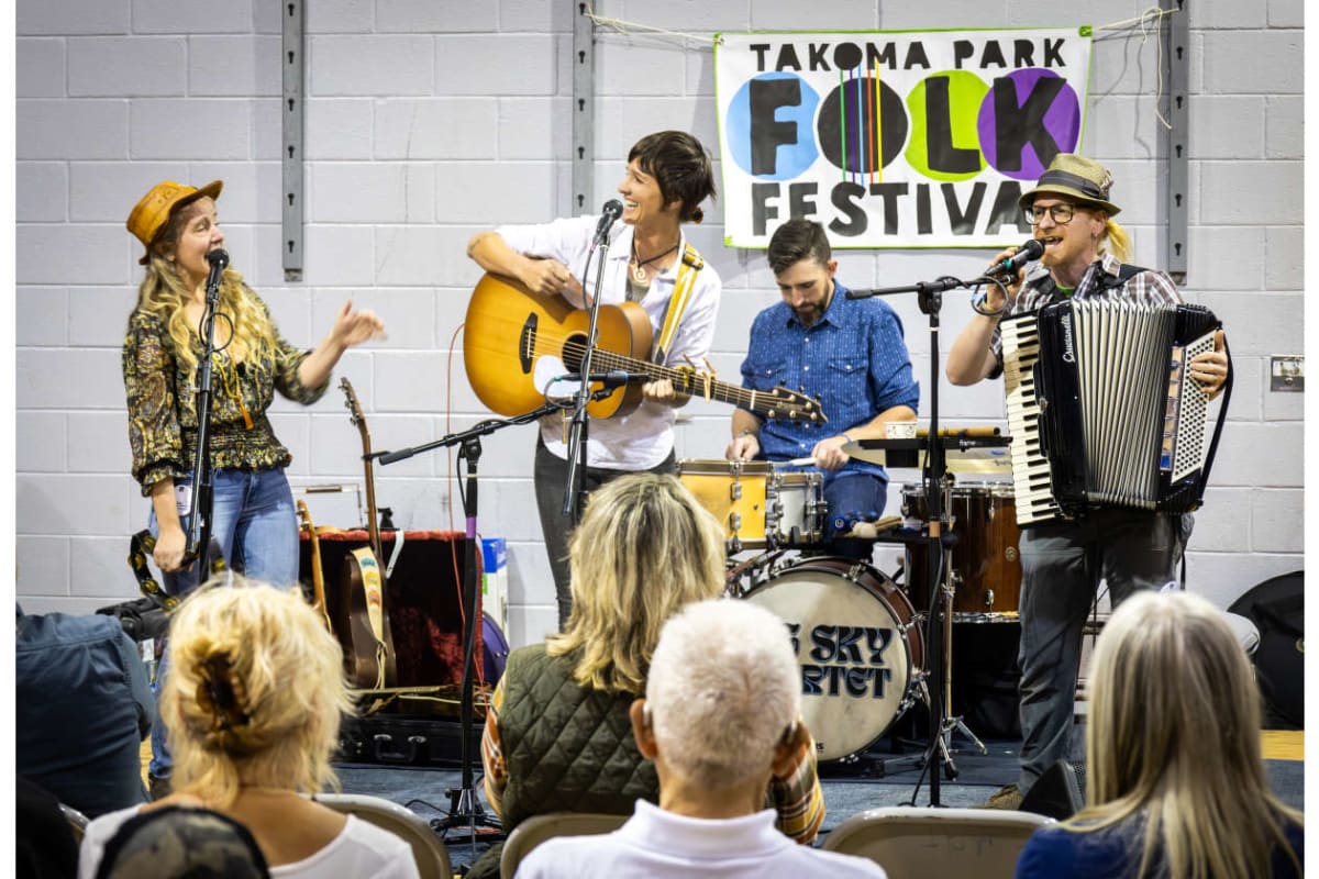 Band performs at Takoma Park Folk Festival; guitarist, singer, drummer, and accordion player on a small stage in front of a banner.