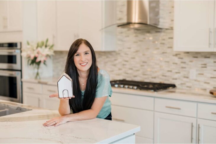 Smiling woman with long dark hair holds a small paper house model in a bright, modern kitchen on a marble island.