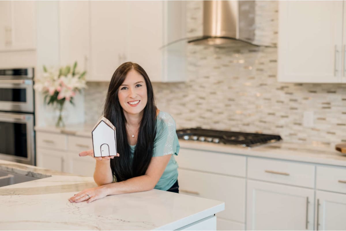 Smiling woman with long dark hair holds a small paper house model in a bright, modern kitchen on a marble island.
