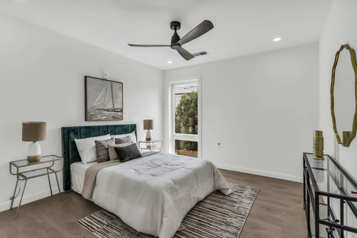 Bedroom with white walls, teal upholstered bed, neutral bedding, two metal nightstands with lamps, sailboat art, and a ceiling fan above.