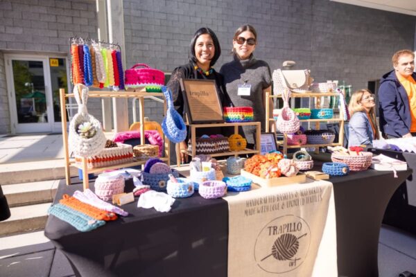 Two smiling women stand behind a market stall displaying colorful crocheted bags, baskets, and accessories in an outdoor setting.