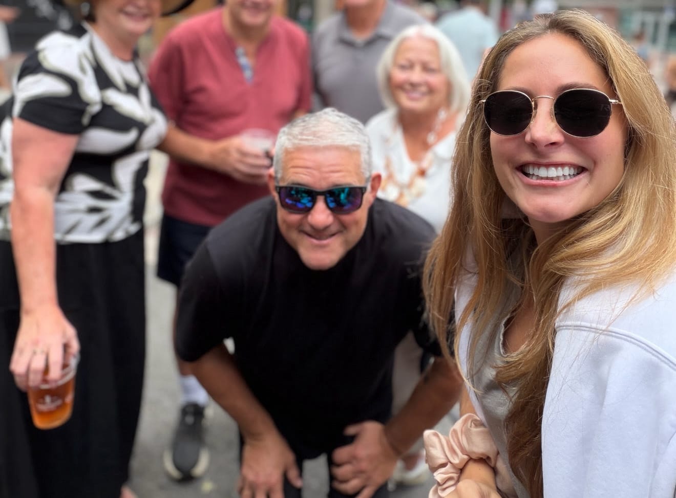 Young woman in sunglasses taking a selfie with a group of friends smiling behind her at an outdoor gathering.