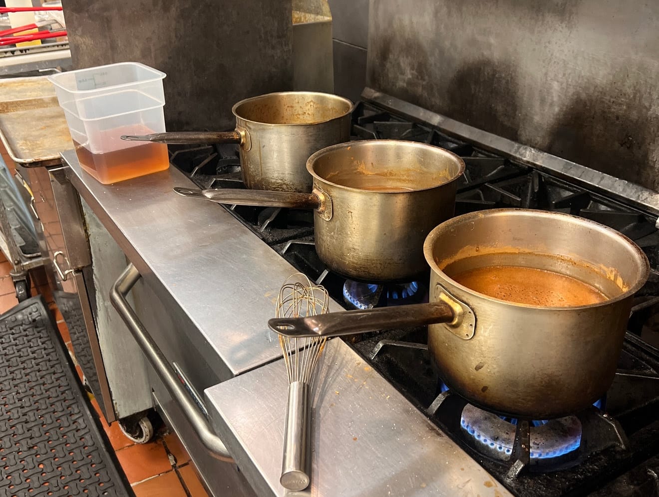 Four stainless pots with brown sauce simmering on a commercial stove; a whisk rests nearby on the prep counter.