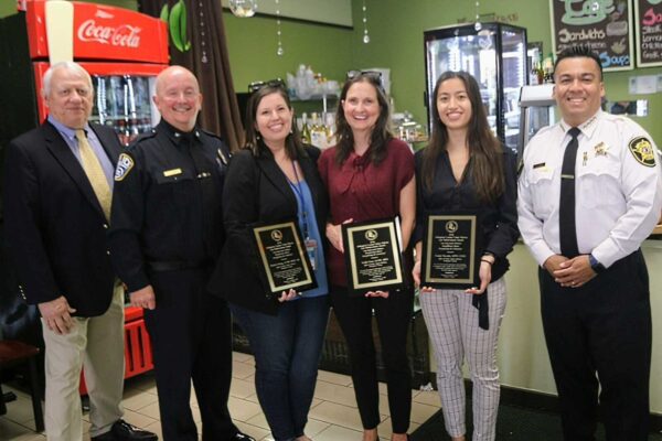 Group of six people in a cafe posing for a photo; three women hold plaques flanked by two uniformed men and a suited man, Coca‑Cola fridge in back.