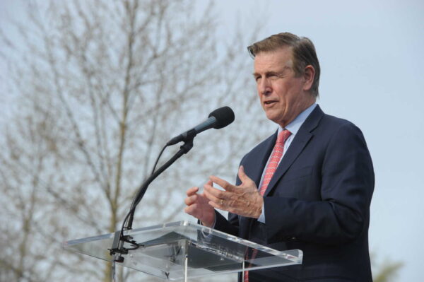 Man in a navy suit and red tie speaks at an outdoor podium with a microphone