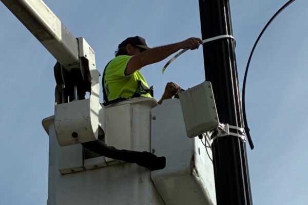Utility worker in a bucket lift securing a cable to a streetlight/pole with tape, wearing a neon vest and cap against a clear blue sky.