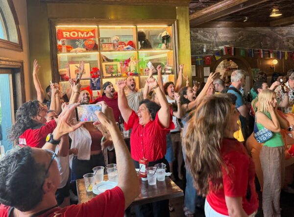 Crowd of people cheering with raised hands at a crowded bar party indoors, some taking photos and wearing red shirts.