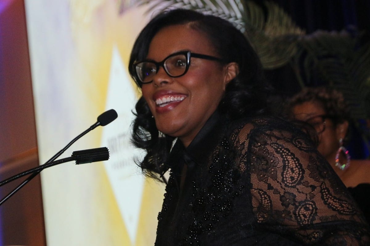 Smiling woman with glasses in a black lace outfit speaks into a microphone at a podium.