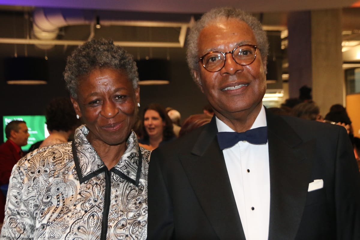 Smiling elderly couple posing at a formal event; man in a tuxedo with bow tie, woman in a patterned blouse beside him.