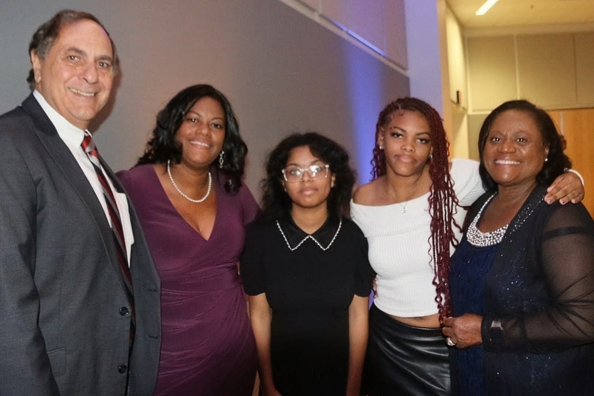 Group of five people posing together at an indoor event, smiling for the camera; man in a suit on the left, others in dresses and smart attire.