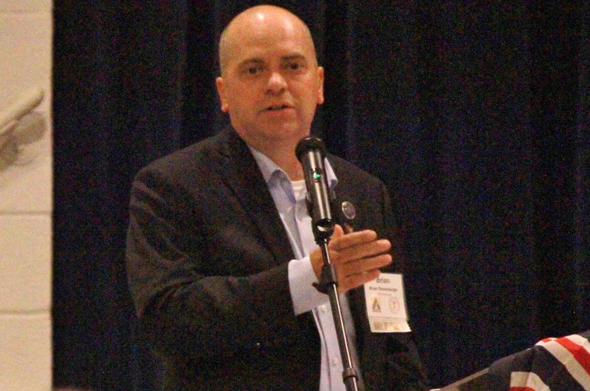 Male speaker in a suit at a microphone on a stage, gesturing with his hands, wearing a name badge.