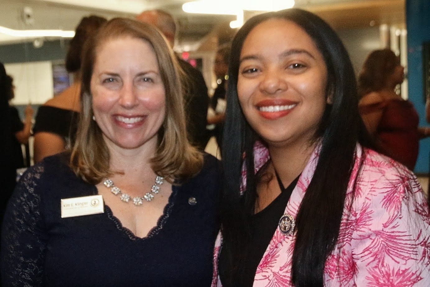 Two women smile for the camera at an event, one wearing a dark lace top with a name badge and statement necklace, the other in a pink patterned jacket.