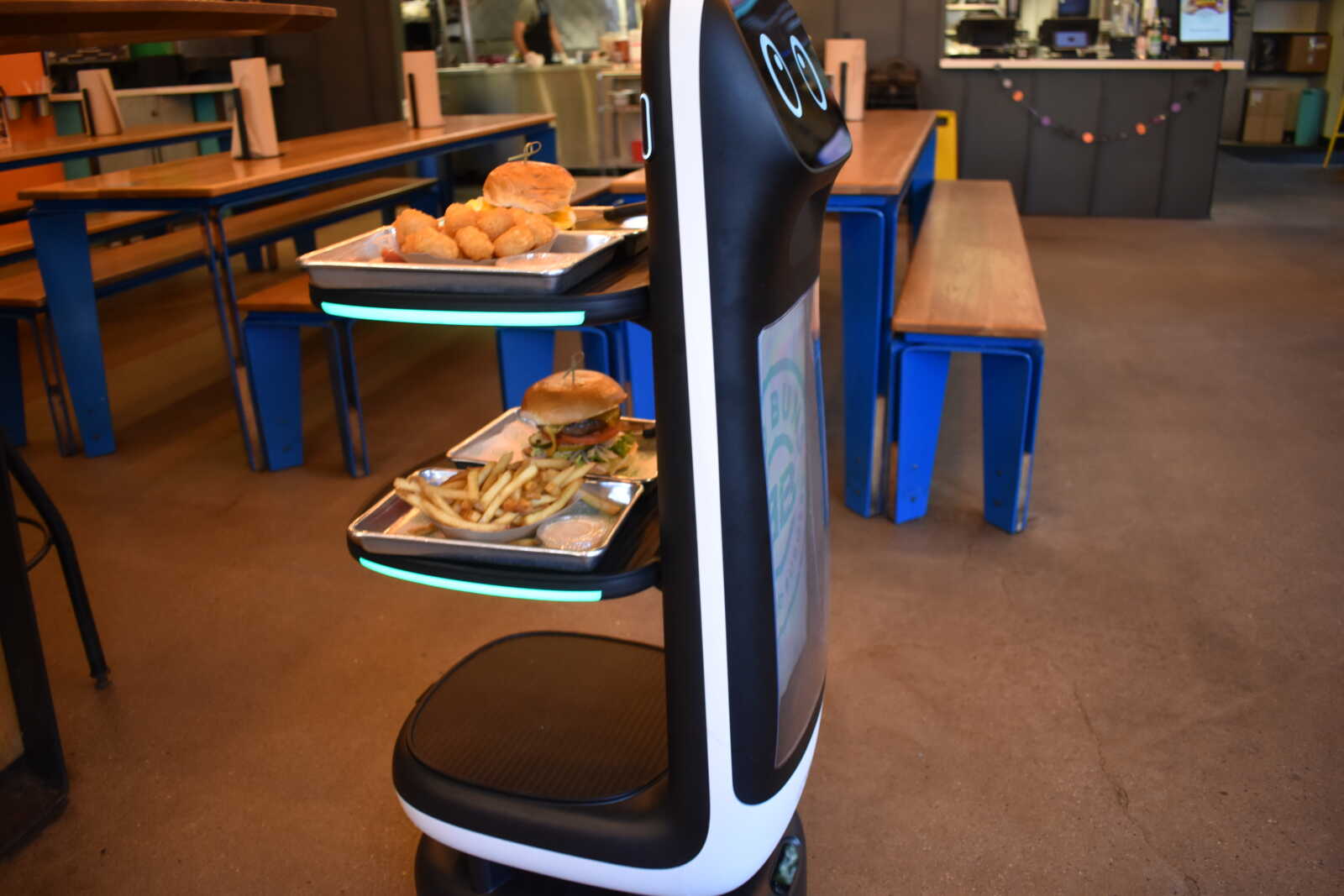 Food-serving robot with trays of burgers, fries, and fried snacks in a cafeteria.