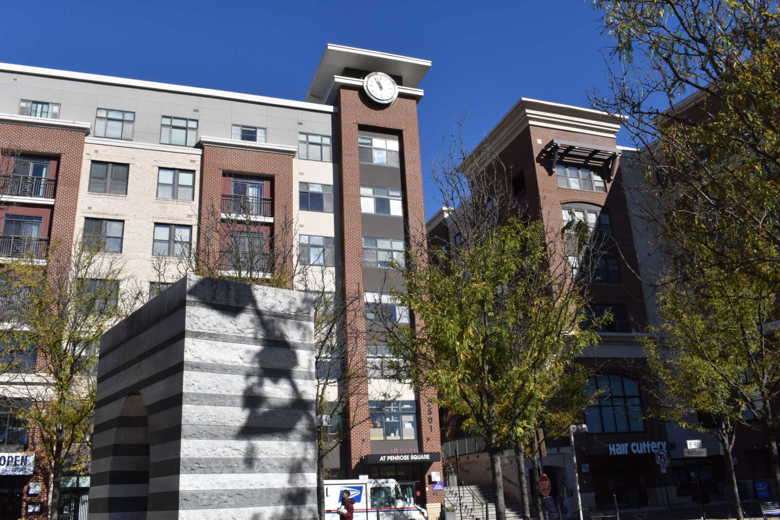 Brick apartment building with a clock tower under a clear blue sky, with trees in the foreground and a striped stone sculpture nearby.