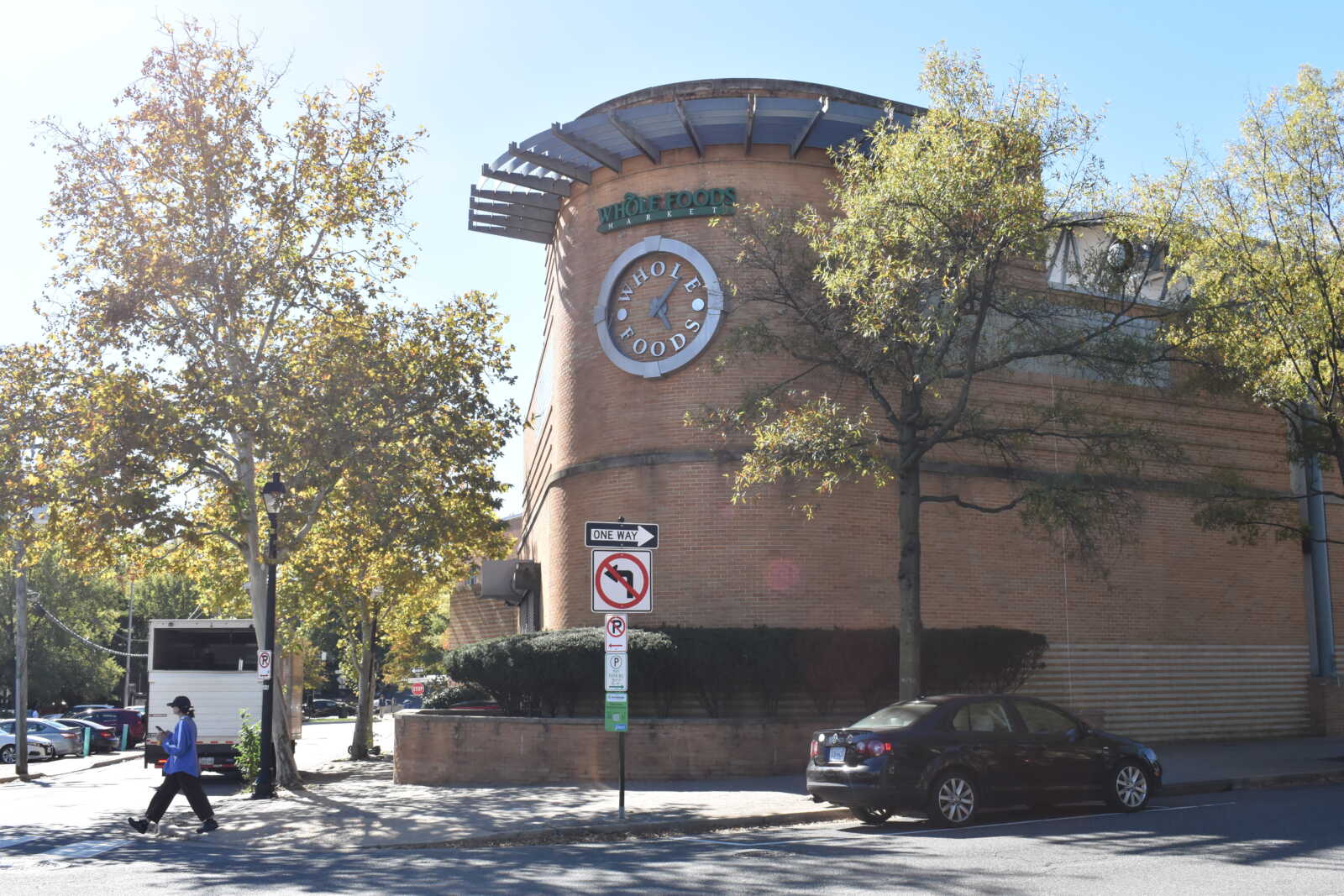 Brick building with a circular Whole Foods Market sign and clock on a tree-lined street; pedestrian crossing in foreground.