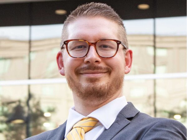 Portrait of a man in a gray suit and yellow striped tie, wearing brown glasses, smiling outdoors in front of glass building windows.