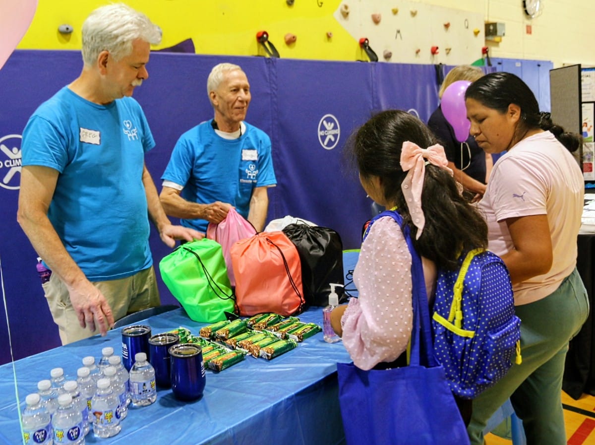 Volunteers in blue shirts hand out snacks and water at a table during a community event in a gym, with kids nearby holding colorful bags.