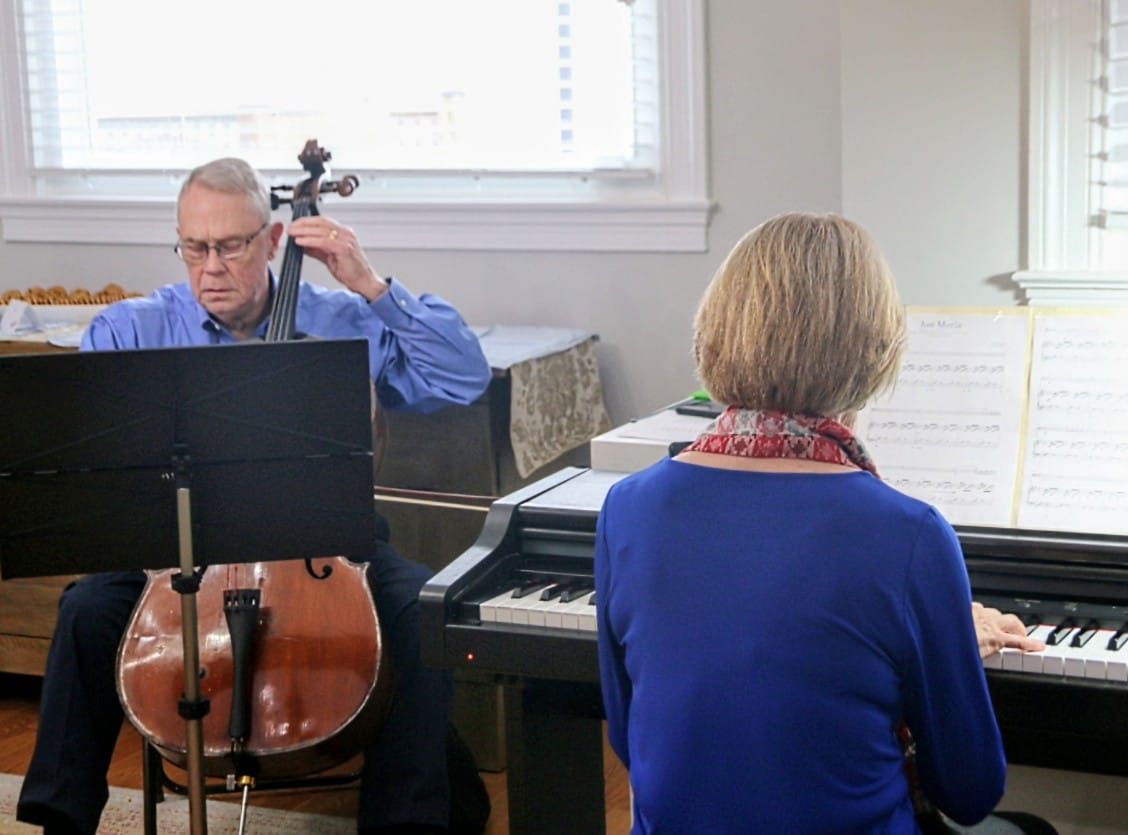 Two musicians—an older man on cello and a woman at a piano—playing in a sunlit room with sheet music on the stand behind them