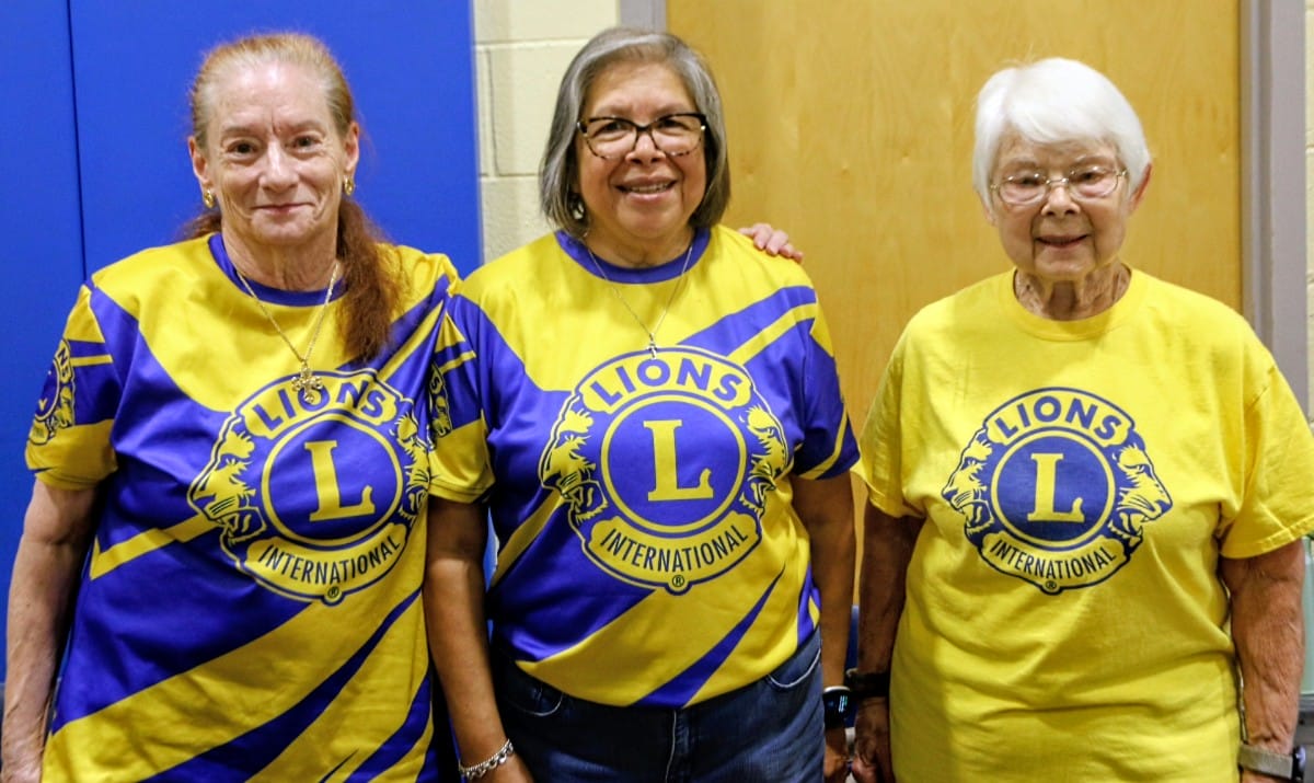 Three women posing together in yellow and blue Lions International shirts, smiling at the camera, logo visible on their shirts.