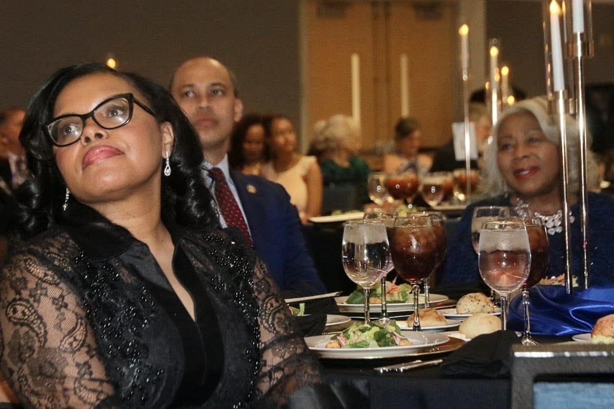 A woman in a black lace dress and glasses sits at a formal banquet table, looking slightly upward as she listens to an event. Behind her are other guests, candles, and plates of food and drinks at the elegant dinner.