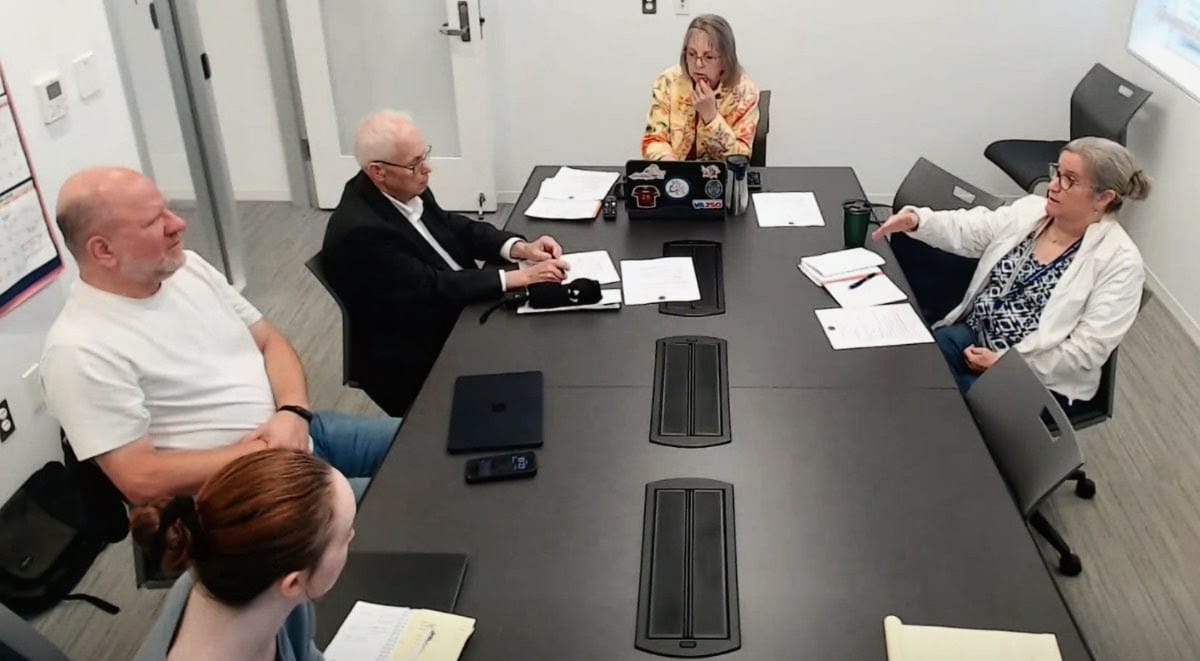 Five people sit around a long conference table in a meeting room, with papers, a notebook, and laptops on the table.