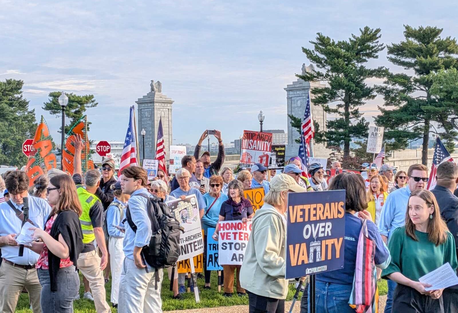 Crowd of protesters in a park holding signs and American flags, with trees and stone arches in the background.