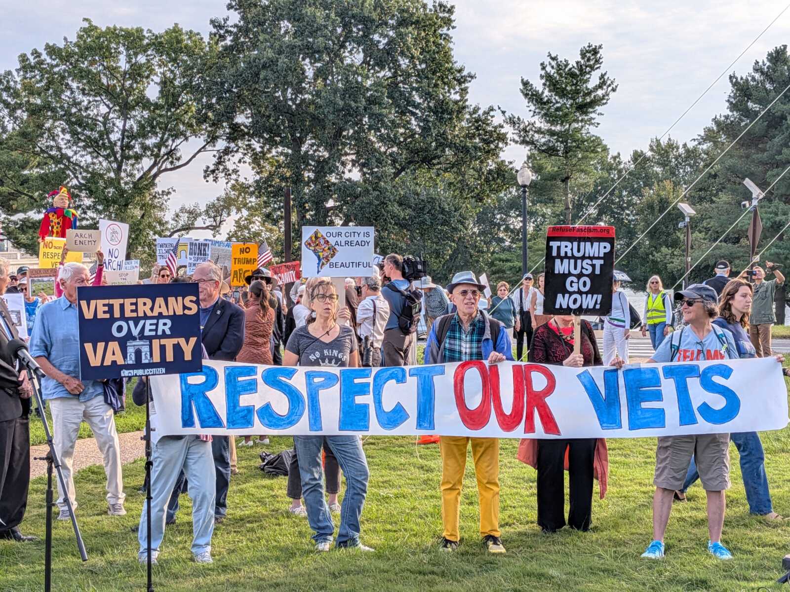 A crowd of protesters holding signs, including a large banner reading 'RESPECT OUR VETS' and a 'VETERANS OVER V A L U T Y' sign, outdoors on a grassy area with trees in the background.