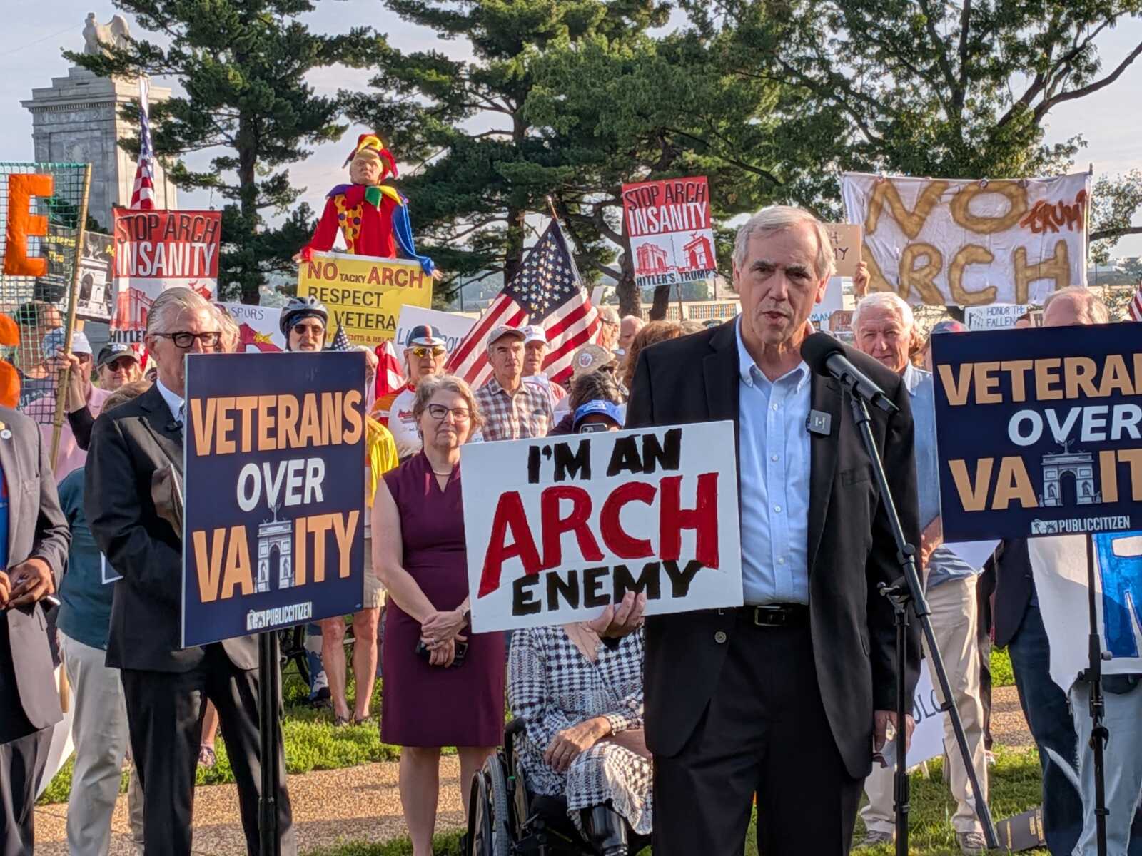 Crowd of protesters holding signs such as 'VETERANS OVER V A L I T Y' and 'I'M AN ARCH ENEMY' with a man at a microphone in front.