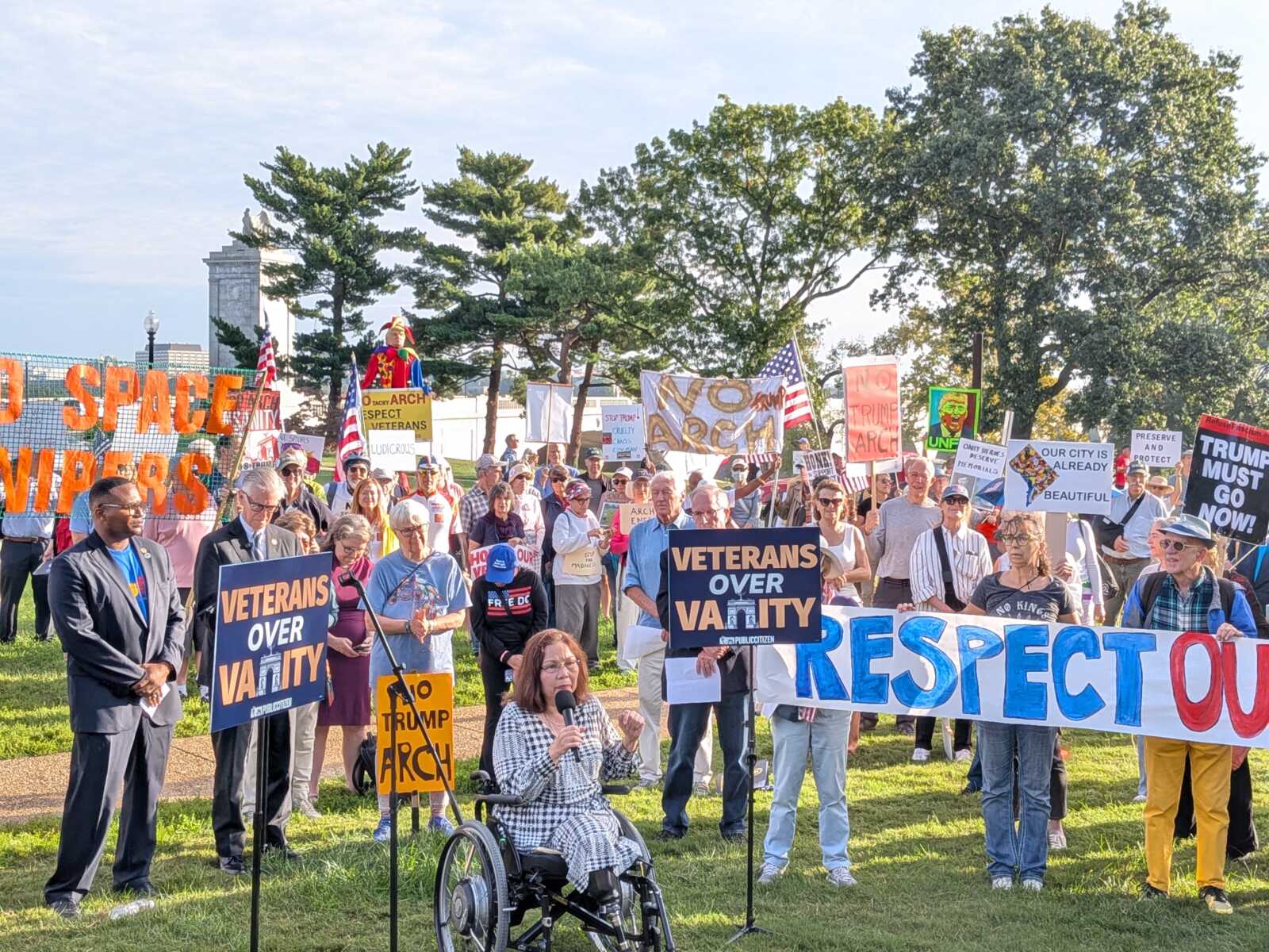 Crowd of protesters outdoors holding colorful signs, including 'VETERANS OVER VANITY' and other banners, with trees in the background.