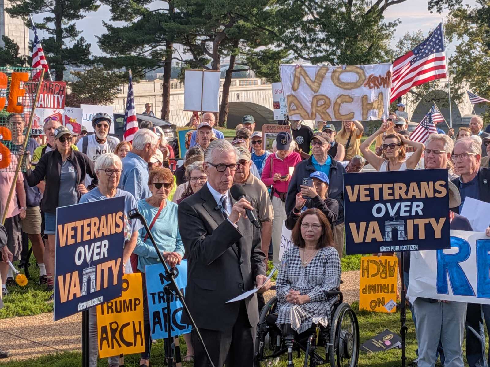Crowd of protesters with signs reading 'VETERANS OVER VANITY' and 'NO ARCH' while a man in a suit speaks into a microphone; American flags waved in background; a person in a wheelchair foreground.
