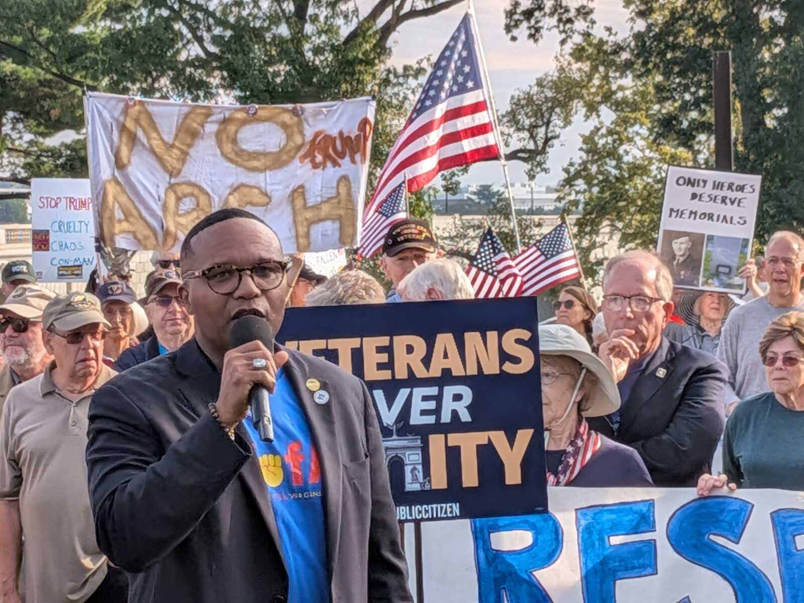 Man wearing glasses and a blue shirt speaks into a microphone at an outdoor protest, with American flags and signs in the crowd behind him.