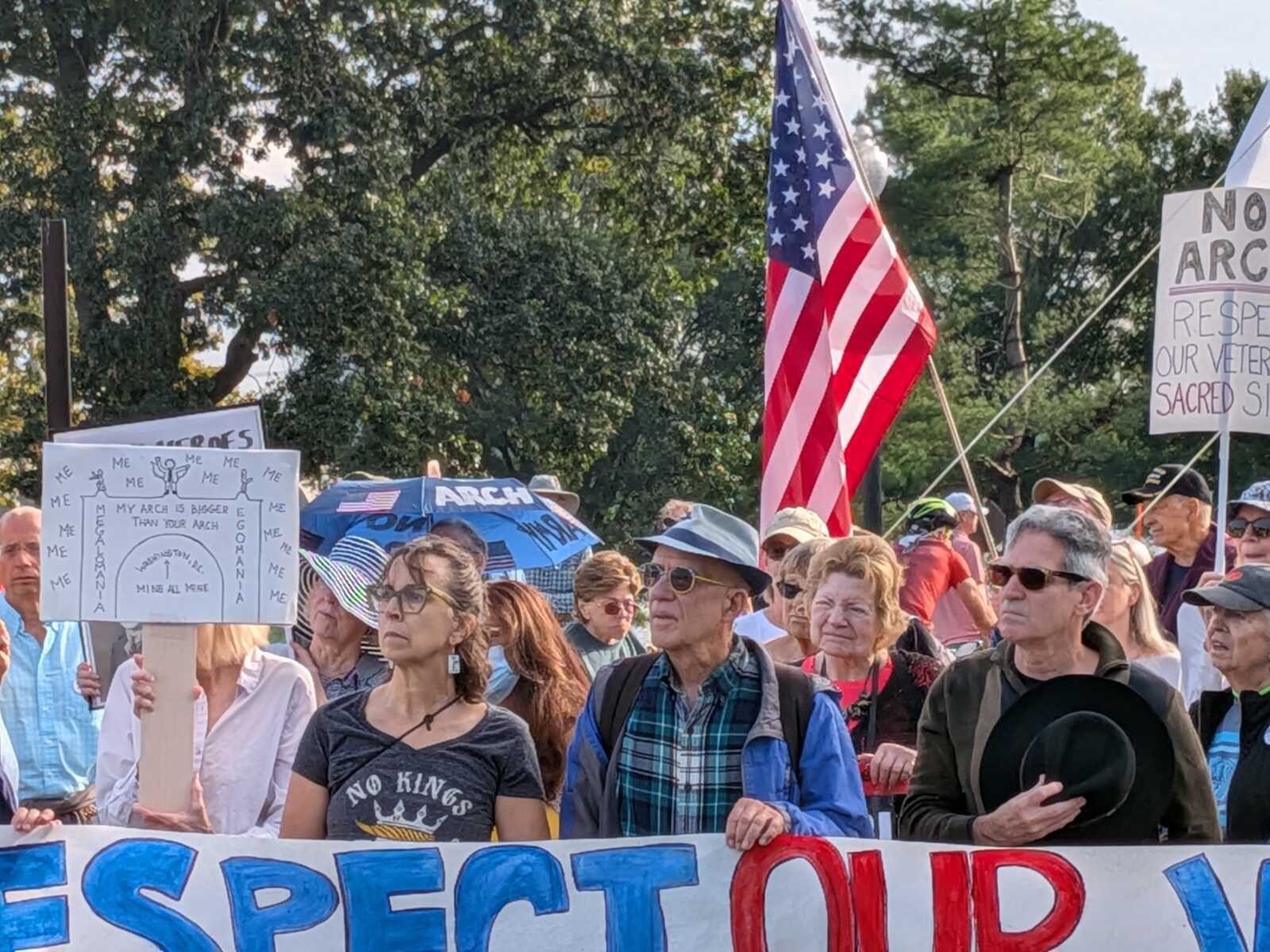 Crowd of protesters at an outdoor demonstration, many holding handmade signs and a waving American flag visible in the center.