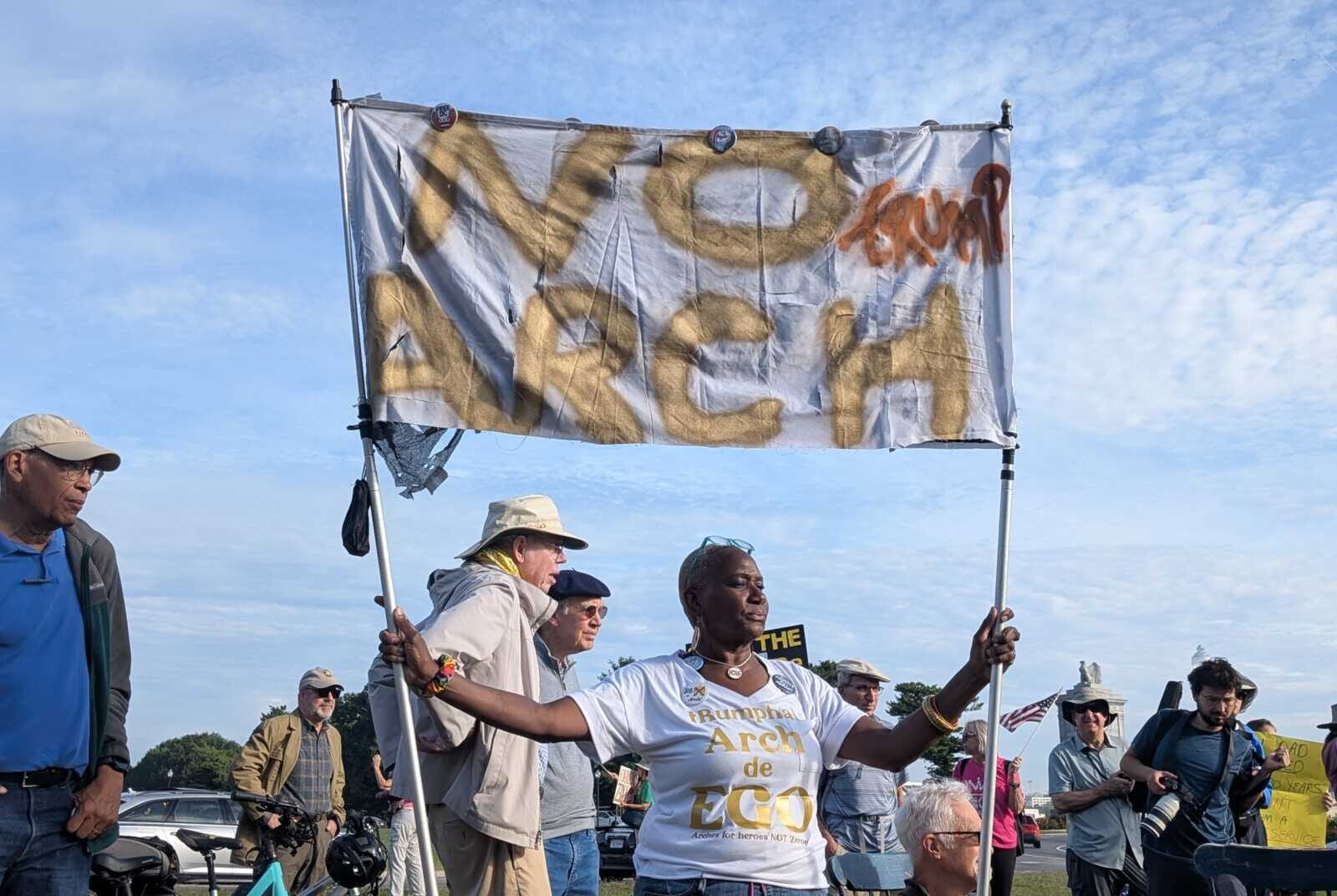 Diverse group of protesters holding a large banner with gold spray-painted letters at a rally, under a clear blue sky.