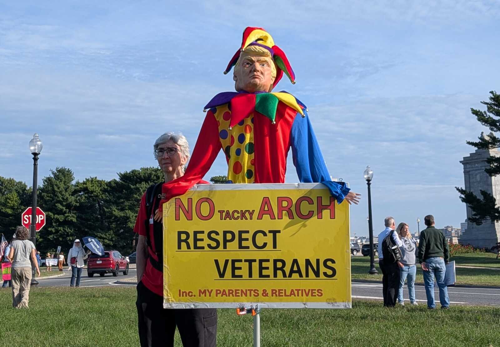 Large colorful clown figure atop a bright yellow protest sign that reads 'NO TACKY ARCH RESPECT VETERANS' with people standing in a park nearby.
