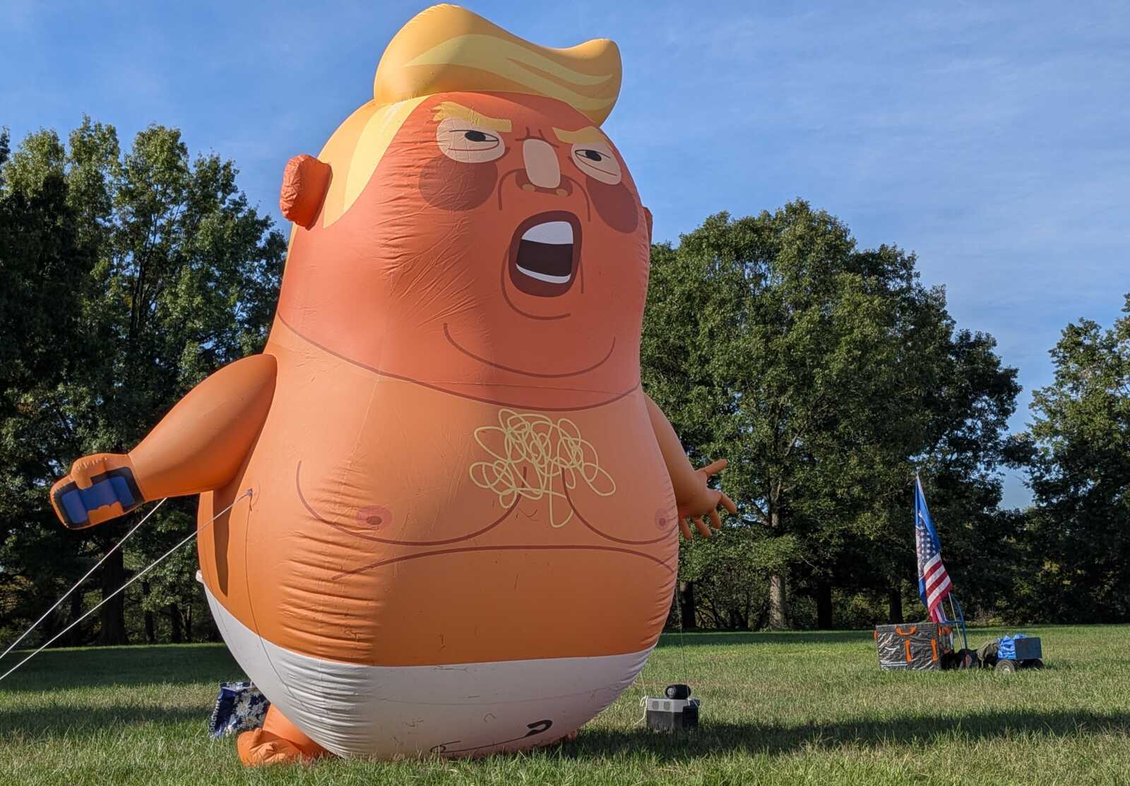 Giant orange inflatable cartoon figure with yellow hair in a park, mouth open, arms out, with an American flag and tents in the background.