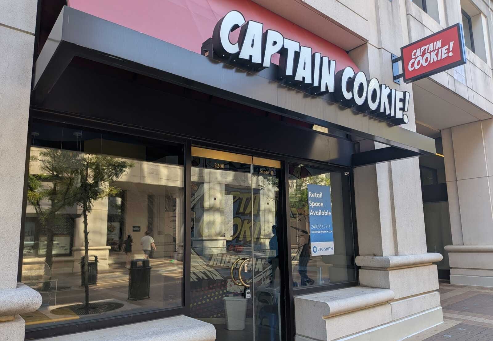 Captain Cookie storefront with a large white logo sign above glass doors and a window display reflecting the street.