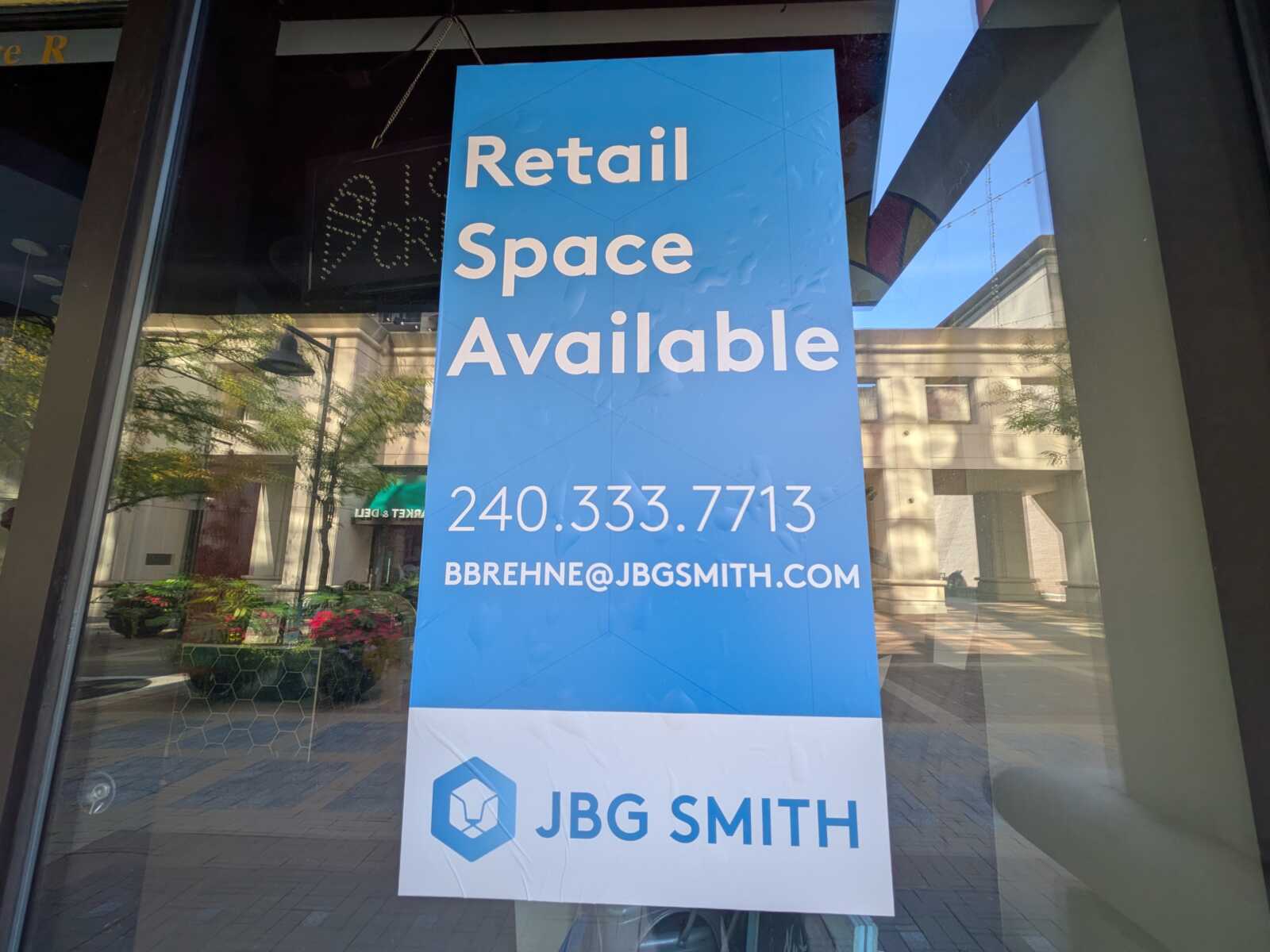 Blue storefront banner reading 'Retail Space Available' with contact 240.333.7713 and email bbrehne@jbgsmith.com, partially reflected in a glass storefront window.