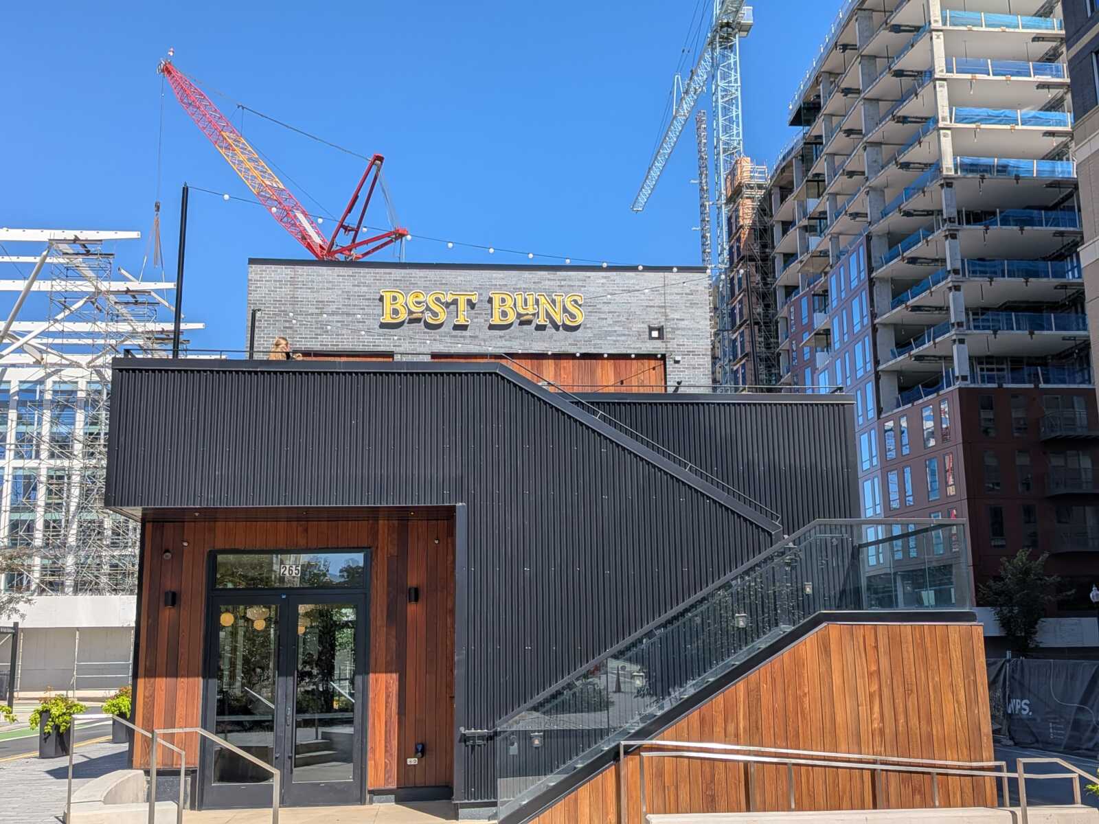 Front of a building with a 'Best Buns' sign, under construction, cranes in the background and blue sky.