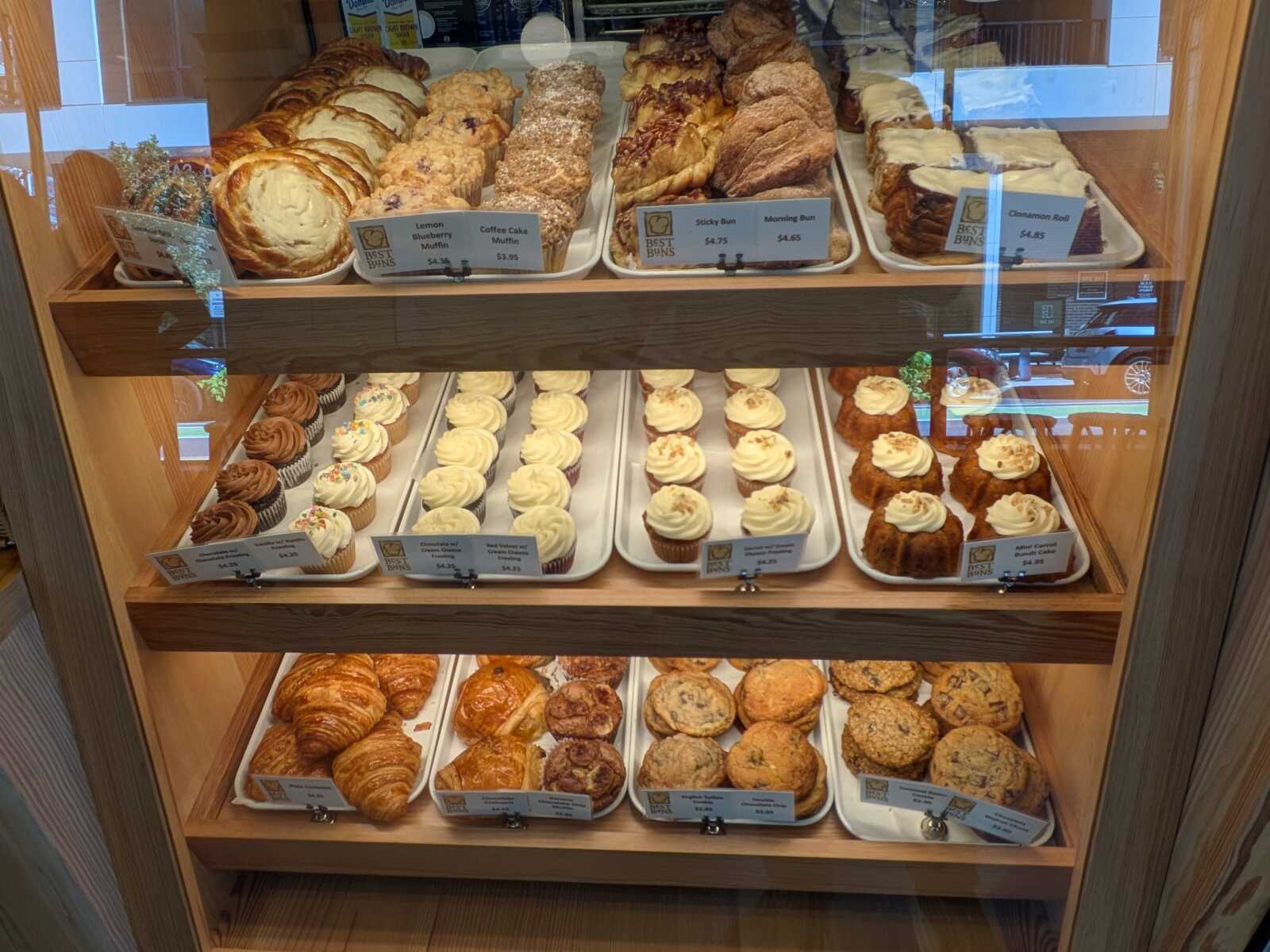 Display case of assorted pastries: croissants, muffins, cinnamon rolls, and cupcakes behind glass.