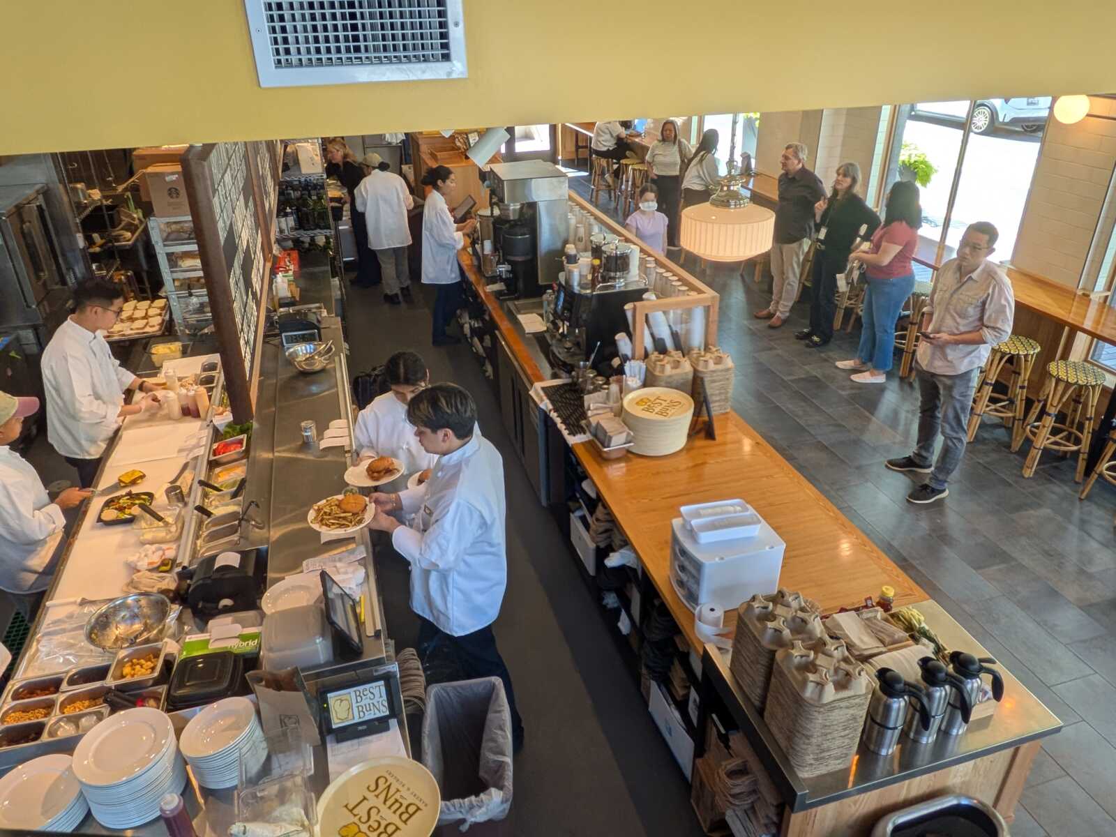 Busy cafe counter scene with chefs serving food and customers in line from an elevated viewpoint