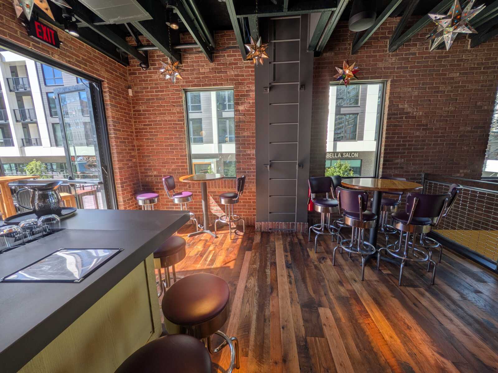Brick-walled cafe interior with round wooden tables, purple stools, and star-shaped lights near large windows showing city storefronts.