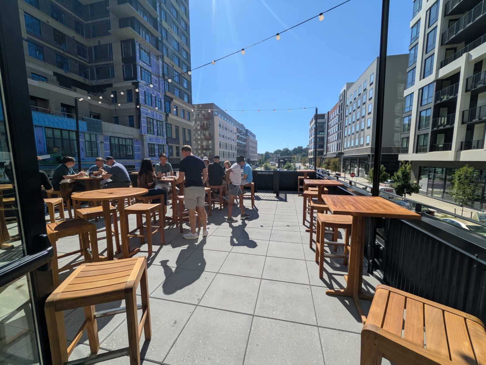 Group of people dining at wooden high-top tables on a sunny rooftop terrace overlooking modern buildings downtown.