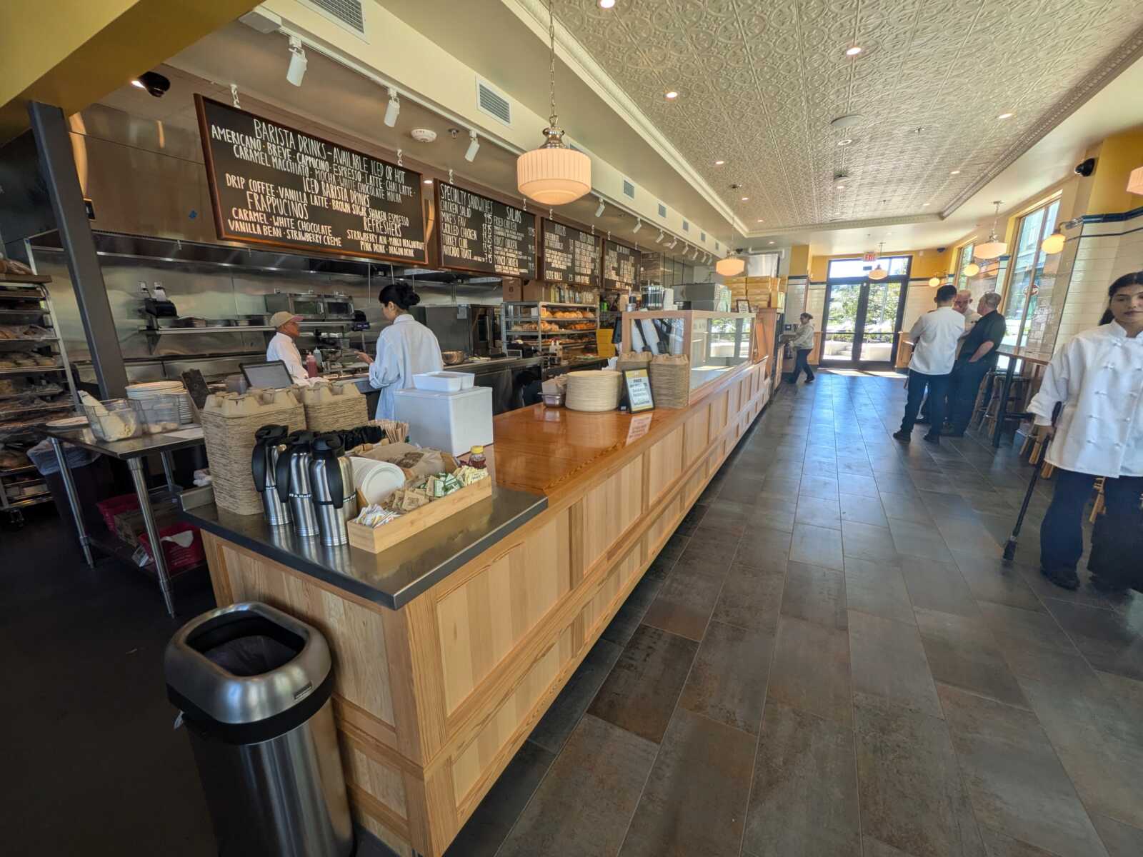 Interior of a cafe with a long wooden counter, chalkboard menus, baristas behind the counter, and customers near the entrance.