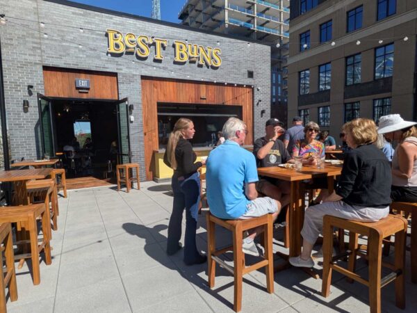Outdoor seating at Best Buns restaurant with people talking and eating at tall wooden tables.