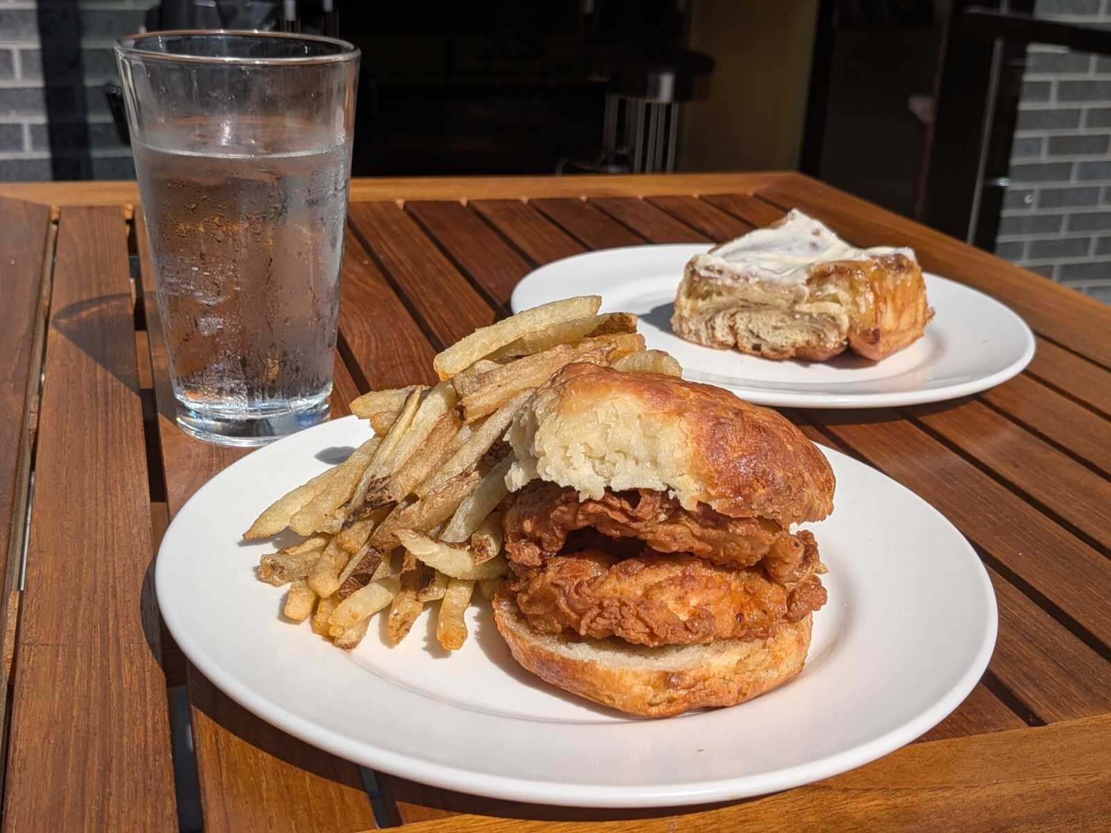 Crispy fried chicken sandwich with a generous stack of French fries on a white plate, beside a tall glass of icy water; a pastry sits on a separate plate in the background on a wooden table.