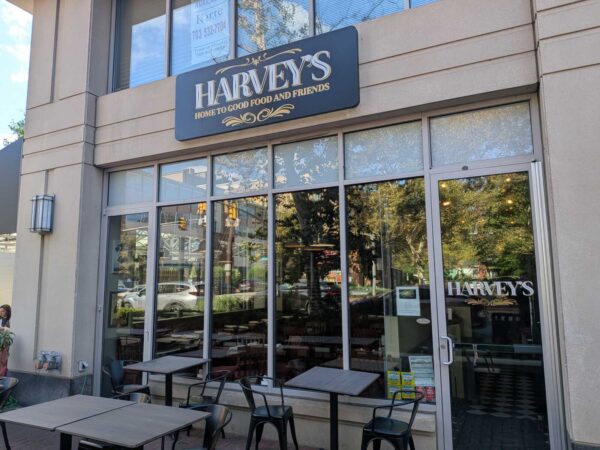 Exterior view of Harvey's restaurant with outdoor seating—tables and metal chairs outside a glass storefront and a blue sign reading 'HARVEY'S HOME TO GOOD FOOD AND FRIENDS'.